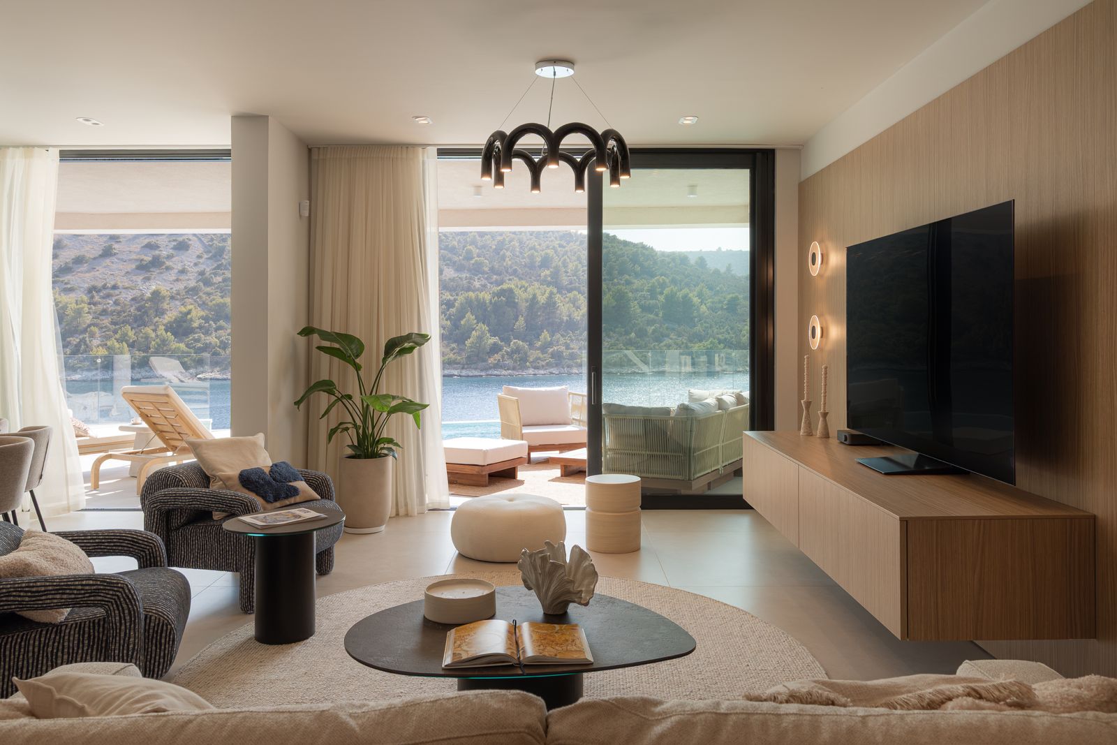 Living room with pale upholstery, timber finishes and full-height glazing opening towards the pool and bay